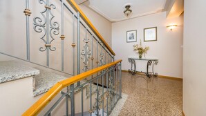 Una escalera con barandillas de metal ornamentadas y un pasamanos de madera, que conduce a un rellano con un cuadro enmarco y una pequeña mesa con un florero.