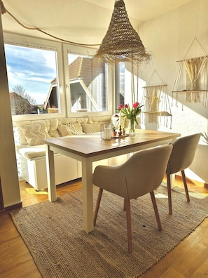 Ein Essbereich mit einem Holztisch und Stühlen, einer gewebten Deckenleuchte und Blick durch große Fenster ins Freie.