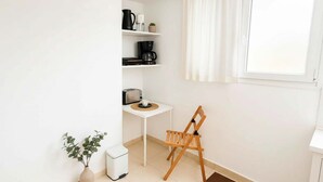 A small, clean room with a white wall, a window with white curtains, a white trash bin, a small table with a cup, a black coffee maker, a toaster, and a wooden easel.