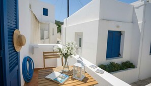 A white building with blue shutters, a wooden chair, a table with a book, a vase with flowers, and a lantern. - Lefkes Home With Village Views (Paros)
