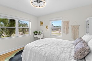 A bedroom with a bed, a desk, a chair, a hanging macrame wall decoration, and a window with a view of palm trees.
