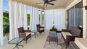 Un patio cubierto con mobiliario de mimbre, un ventilador de techo y vistas al verdor exterior.