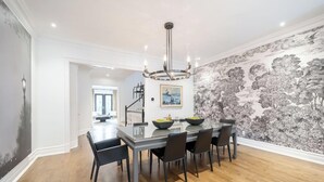 A dining area with a glass table, black chairs, a chandelier, and a mural with trees. - Rare Yorkville Detached House with Parking and Hot Tub (Toronto)