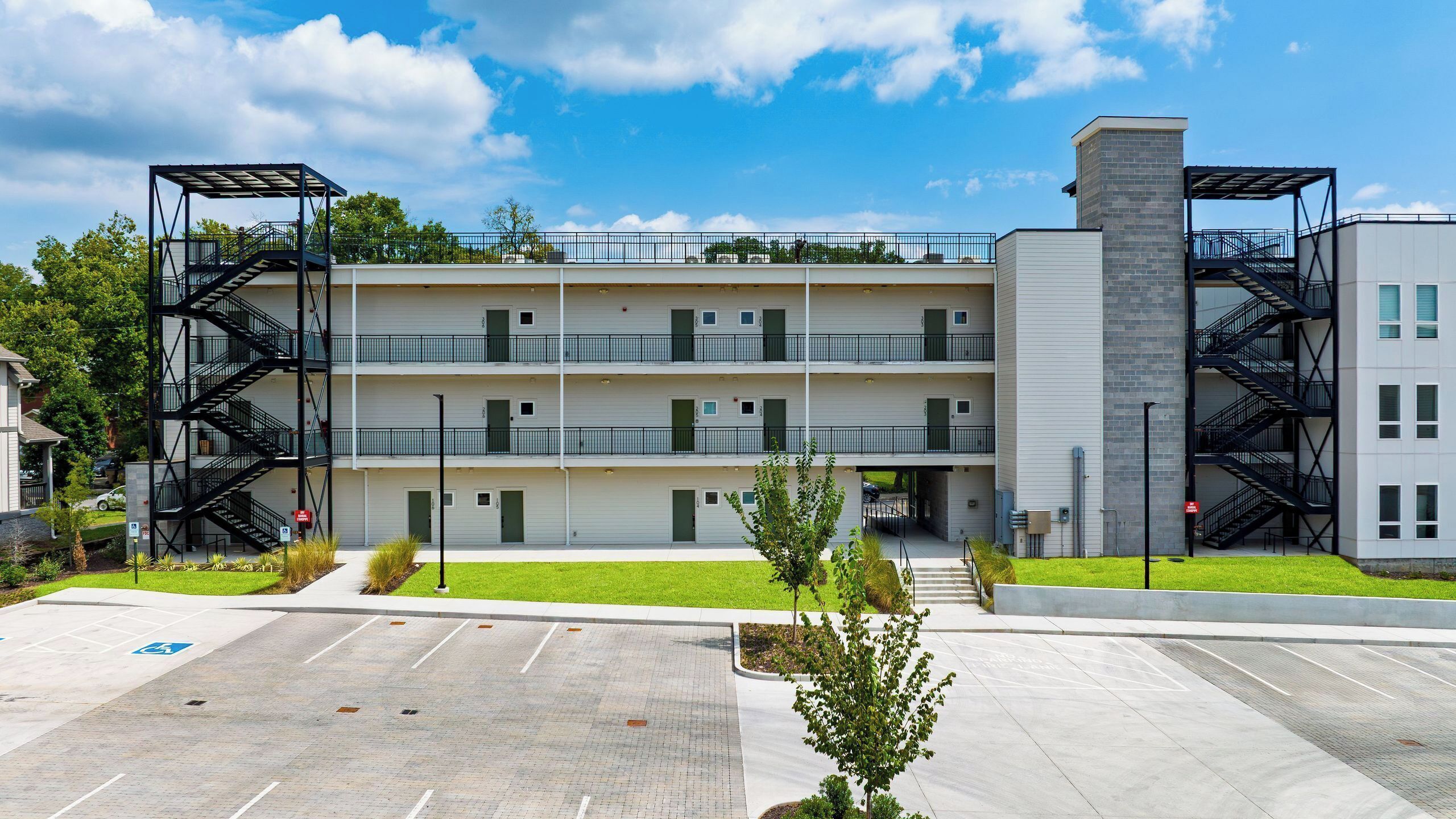 A multi-story building with external fire escapes, a parking lot, and a small green area with trees.
