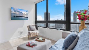 A modern living room with a large window, a view of the city skyline, a cozy seating area, and a small table with decorative items. - Modern Downtown Suite with Full Amenity Access (Miami)