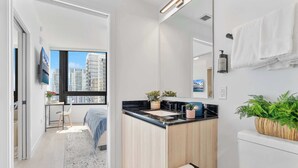 A modern bathroom with a large mirror, a sink, and a toilet. There is a window with a view of buildings and a door leading to a bedroom. - Modern Downtown Suite with Full Amenity Access (Miami)