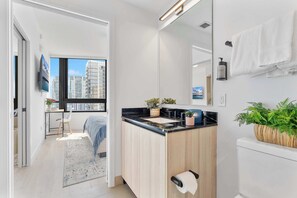 A modern bathroom with a large mirror, a sink, and a toilet. There is a window with a view of buildings and a door leading to a bedroom. - Modern Downtown Suite | Walk to Bayfront Park (Miami)