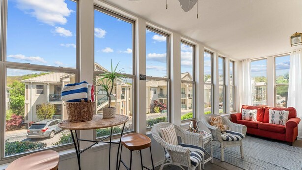 A sunlit room with large windows, wicker furniture, a red sofa, and a view of neighboring houses. - Emerald Bay Escape with Sunroom (Hollister)