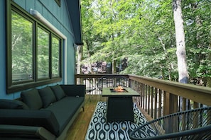 A deck with a sofa, a table with a fire pit, and a view of a forest. - Cozy 4-bedroom chalet in The Hideout pet friendly with AC, hot tub (Cobbs Lake Preserve)