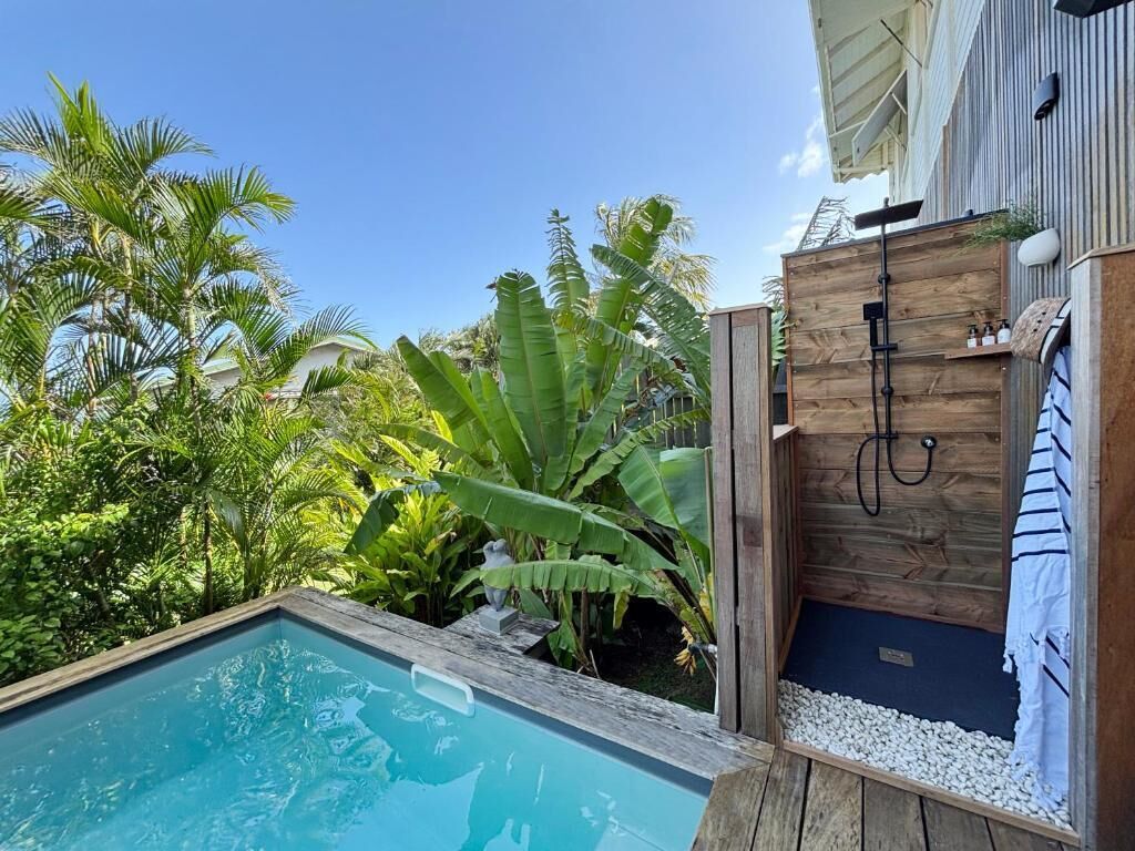 Une piscine dotée d’une terrasse en bois, entourée d’une végétation luxuriante et bordée d’un mur en bois sur lequel est accrochée une serviette.