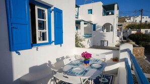 A white building with blue shutters and a table set for two with a vase of flowers. - Ornos Home With Sea View (Mykonos)