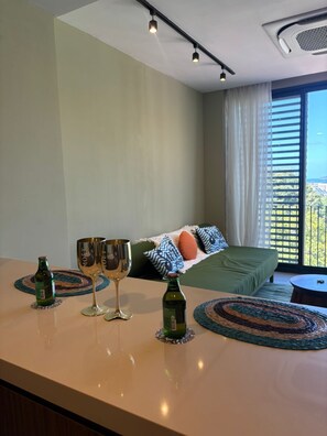 Una camera da letto moderna con un letto, un comodino con due bicchieri e bottiglie di vino, e vista sul verde dalla finestra.