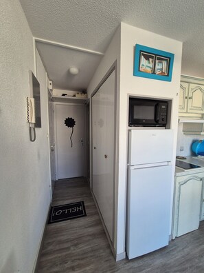 A narrow hallway leading to a kitchen with a white refrigerator, a microwave, and a blue-framed picture above the microwave. - Apartment T2 Centre Port Cap d'Agde (Agde)