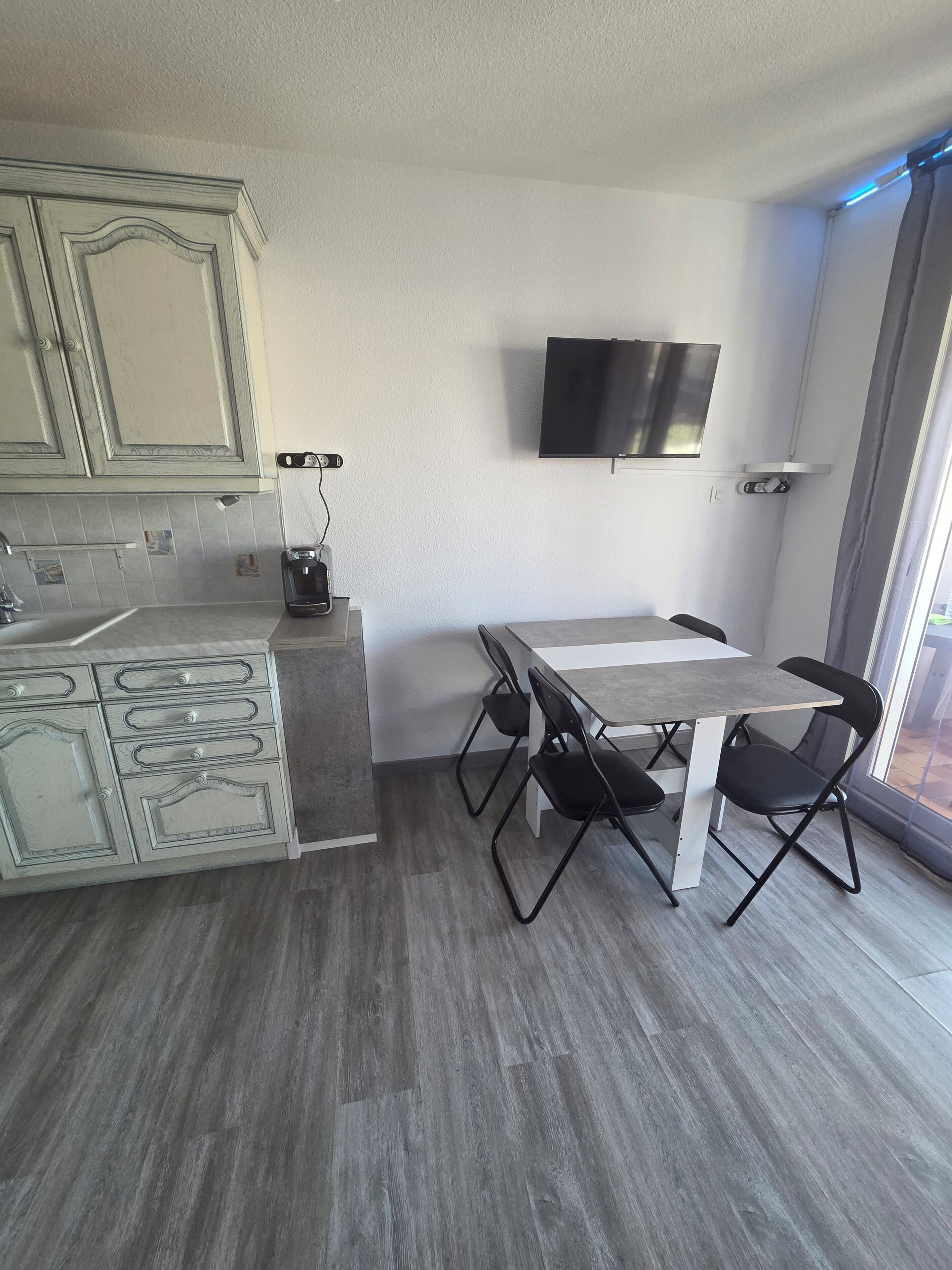 Une cuisine moderne avec un coin repas, un téléviseur à écran plat fixé au mur et une grande fenêtre laissant entrer la lumière naturelle.