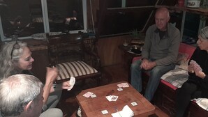 A group of people playing cards around a wooden table in a room with wooden furniture and a window. - Hostal La Minga (Chaiten)