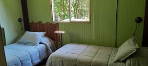 A room with two beds, a small round table, a window with a view of trees, and a wooden headboard. - Hostal La Minga (Chaiten)