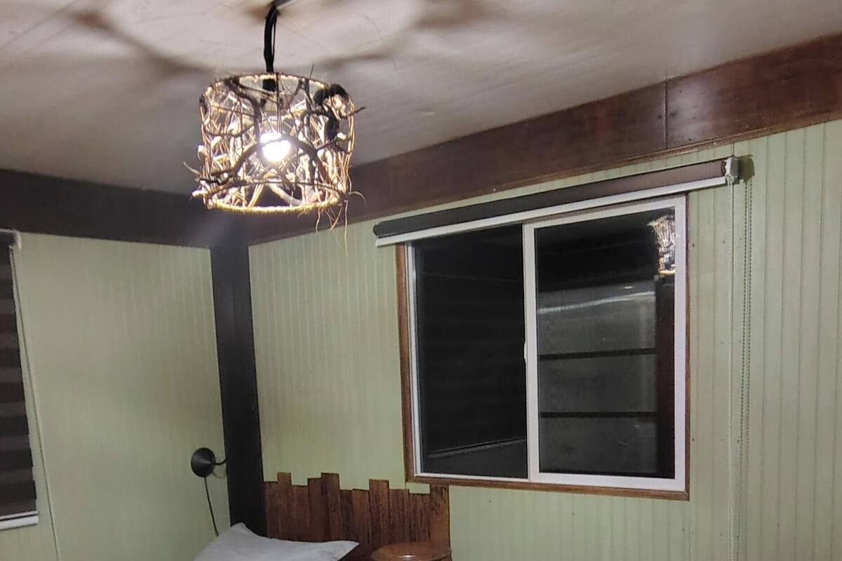 Ein Zimmer mit zwei Betten, Holzboden, einem Fenster mit Jalousien und einer Hängeleuchte.