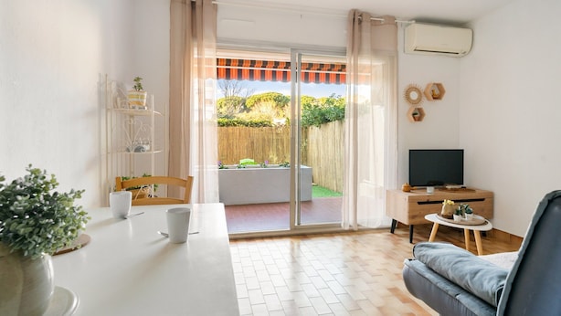 A living room with a sofa, a TV on a stand, a dining table with chairs, and a sliding glass door leading to an outdoor area with a wooden fence and greenery. - Carry Le Rouet, Proche Plages, Commerces (Carry-le-Rouet)