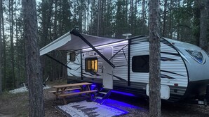 En autocamper med et overdækket udendørs område, et picnicbord og en blå oplyst måtte.