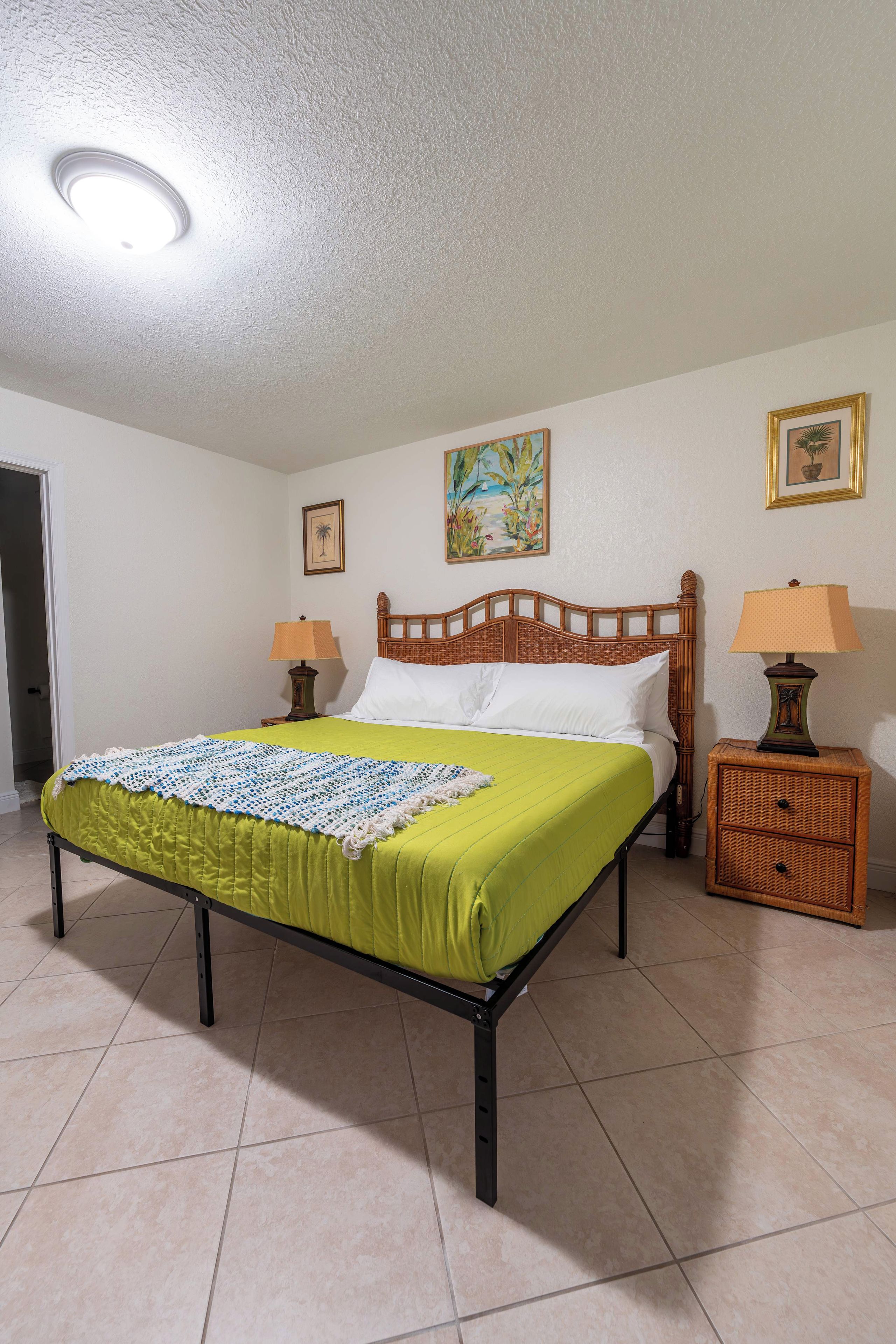 A single-bed room with a wooden headboard, a green bedspread, two bedside tables with lamps, and a door to the left.