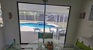 Una mesa de vidrio con un plato de condimentos, una puerta corrediza de vidrio que da a un patio trasero con piscina y mobiliario de exterior.