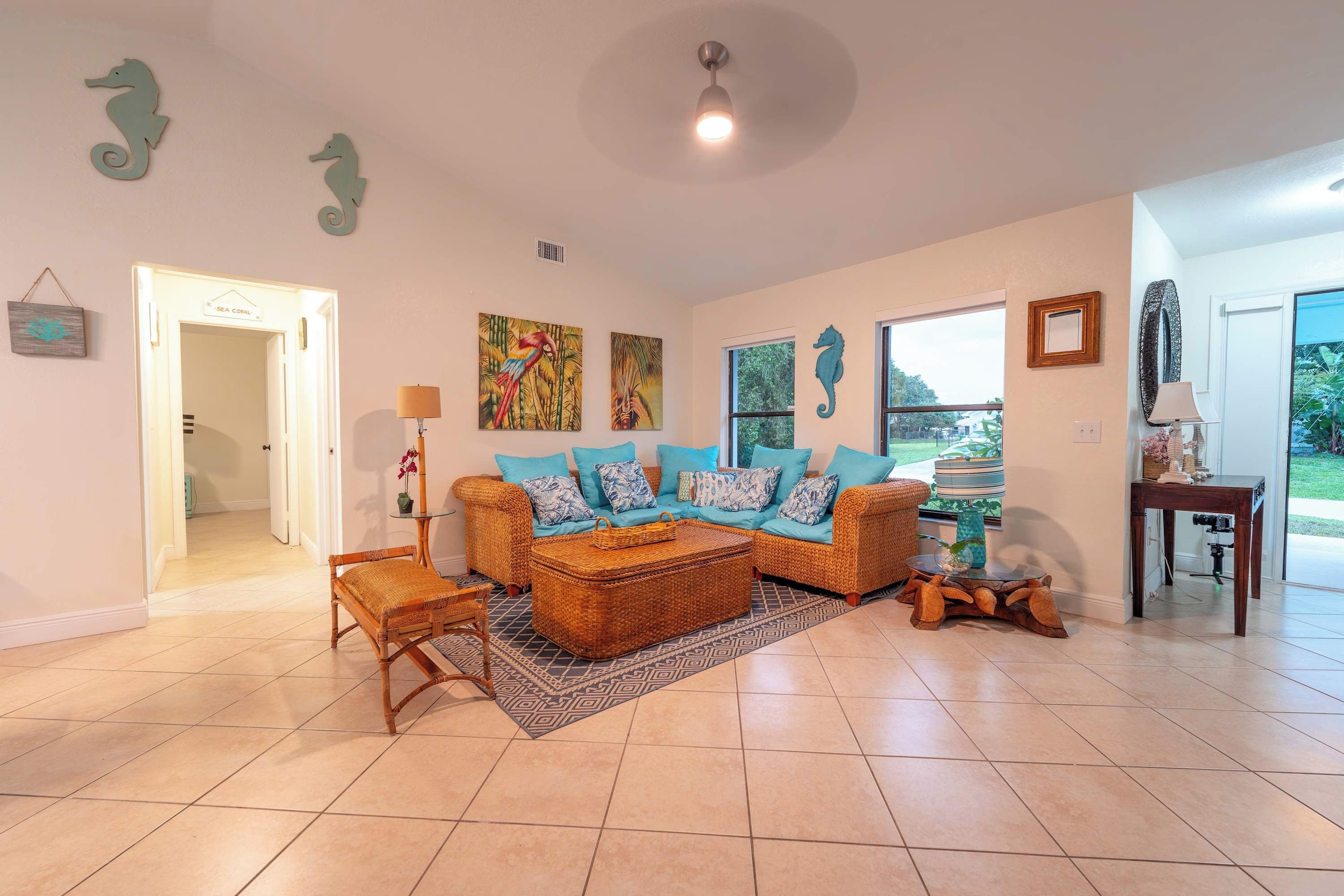 A living room with a sofa set, a coffee table, and a rug. There are two seahorse wall decorations, a painting of a parrot, and a window with a view of greenery.