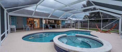 A covered outdoor swimming pool area with a hot tub, seating, and a view of the house.
