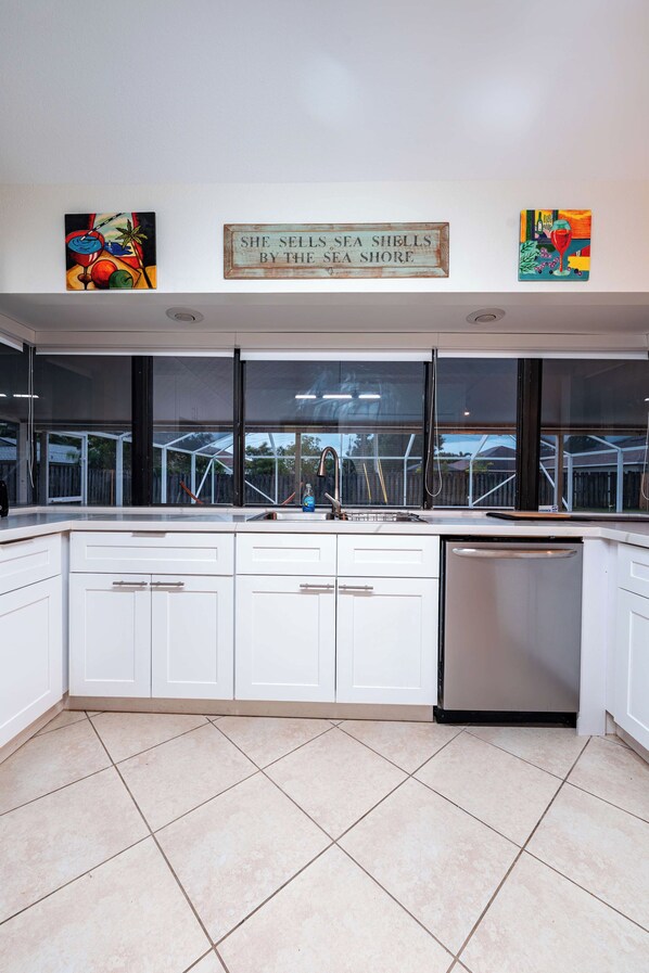 A modern kitchen with white cabinets, a dishwasher, and a large window overlooking a backyard.