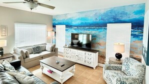 Uma sala de estar com ventilador de teto, uma televisão grande e um mural de praia na parede.