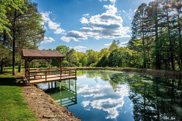 A wooden gazebo over a calm pond, surrounded by lush greenery and tall trees.