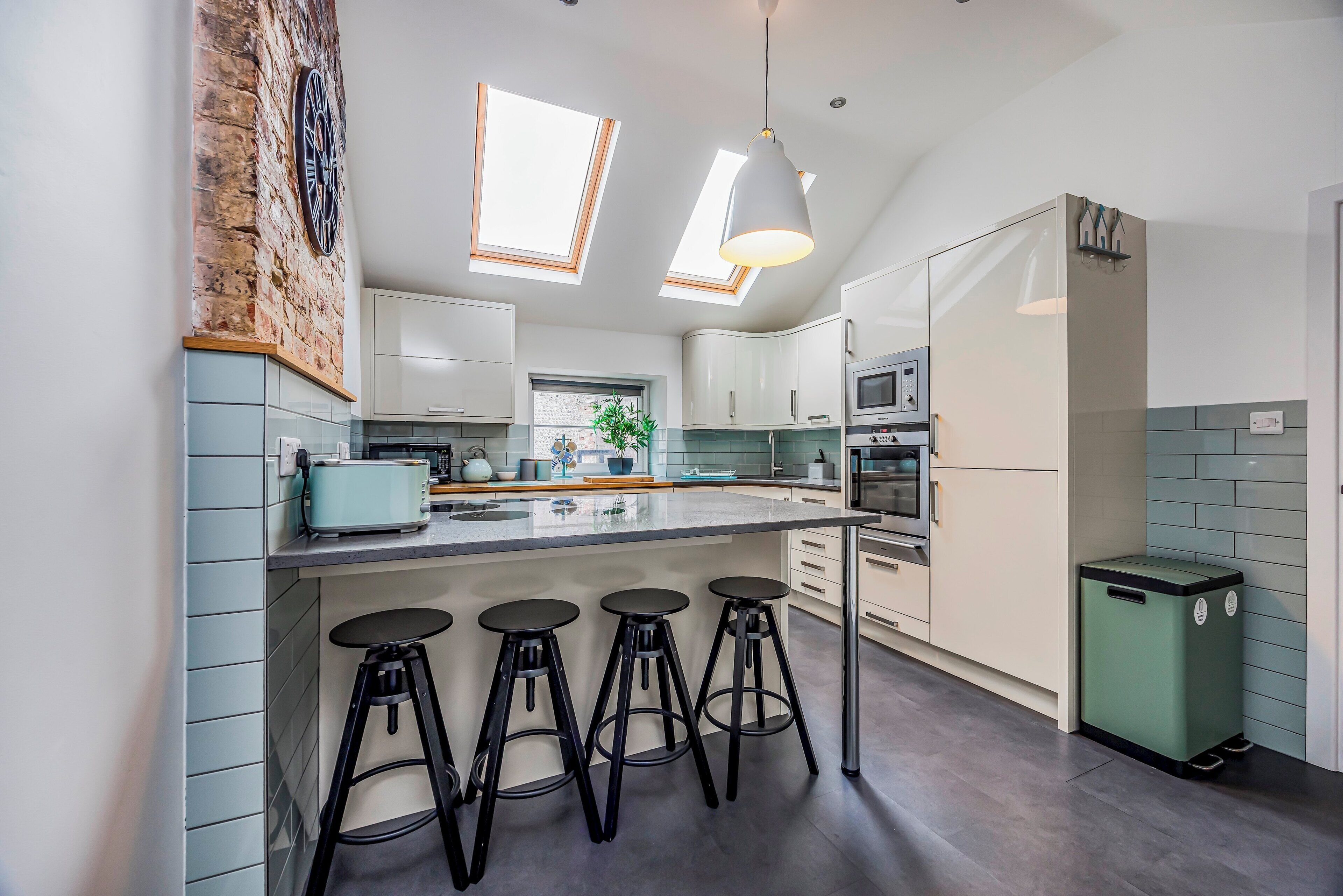 A modern kitchen with a central island, bar stools, skylights, and a green trash bin.
