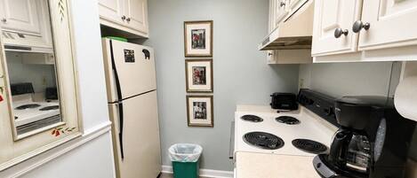 A compact kitchen with white cabinets, a stove, a refrigerator, and a trash bin.