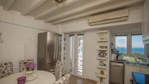 Uma cozinha moderna com geladeira de aço inoxidável, sala de jantar com mesa redonda e cadeiras florais, e vista para o mar através de grandes janelas.