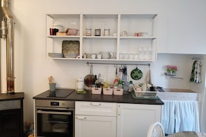 Une cuisine équipée d’une cuisinière, d’un four et de meubles blancs. Des étagères au-dessus du plan de travail servent à ranger la vaisselle et les bocaux.