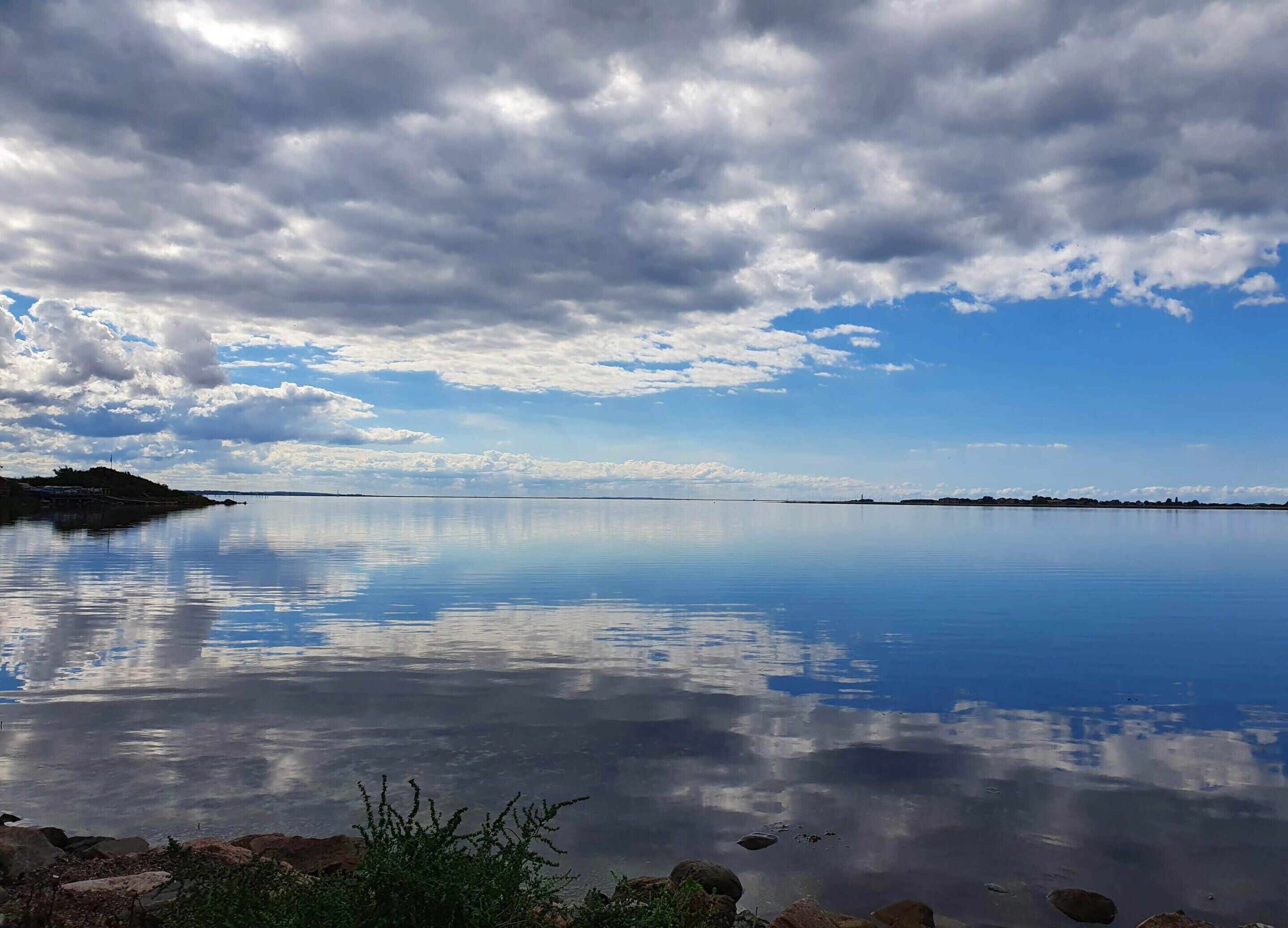 A calm lake with a clear reflection of the sky and clouds, surrounded by rocky terrain and sparse vegetation.