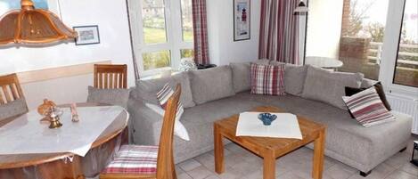 Un salon chaleureux avec un canapé, une table à manger, des chaises et une table basse. Il y a une fenêtre avec des rideaux, un luminaire suspendu et un tableau encadré au mur.