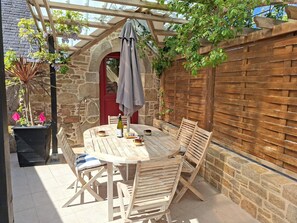 Una mesa y sillas de madera bajo una pérgola de madera, con un muro de piedra y una puerta roja al fondo.