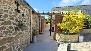 Un edificio de piedra con una pérgola de madera, plantas en macetas y un camino empedrado.