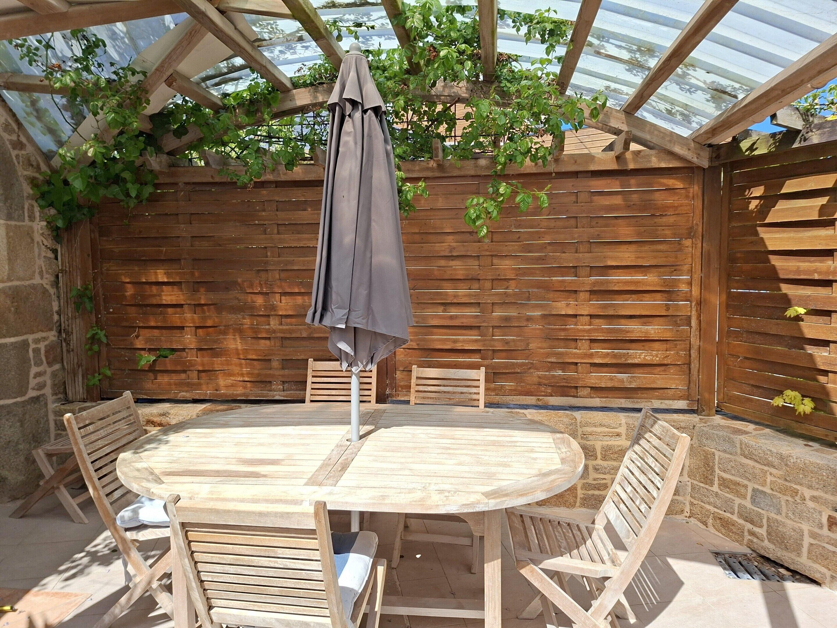 Una pergola in legno con tavolo e sedie, un ombrellone grigio e piante rampicanti.