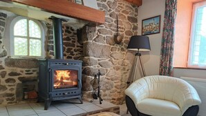 Una habitación con paredes de piedra, una estufa de leña, un sillón acolchado y suelo de baldosas.