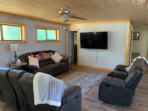 Ein Wohnzimmer mit einer Couch, zwei Sesseln, einem an der Wand befestigten Fernseher, einem Deckenventilator und großen Fenstern.