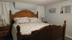 Une chambre à coucher avec un lit en bois, une table de chevet, une lampe, un tableau encadré au mur et une fenêtre avec des rideaux.