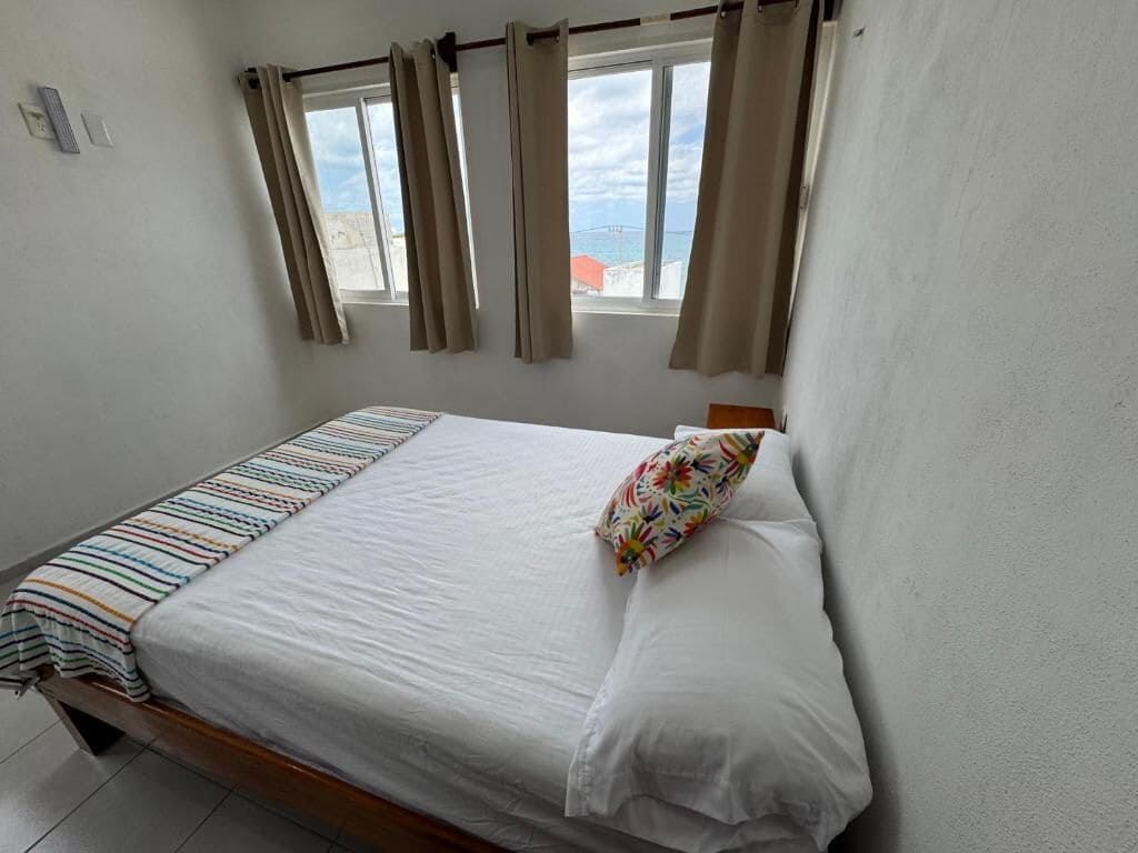 A single bed with a striped blanket, a colorful pillow, and a window with curtains.