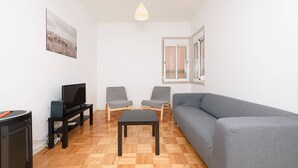 Ein Wohnzimmer mit grauem Sofa, zwei grauen Sesseln, einer schwarzen TV-Konsole, einem schwarzen Tisch und einer Pendelleuchte.