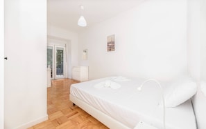 Ein Schlafzimmer mit Bett, Nachttisch, Lampe, Fenster mit Blick und einer Tür, die auf einen Balkon führt.