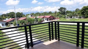 A balcony with a black metal railing, a wooden floor, and a view of houses and greenery in the background. - Cozy Corner Transient House (Calapan City)