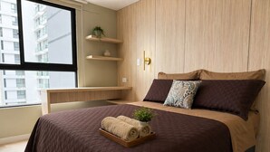 A modern bedroom with a large bed, a wooden headboard, a small desk, and a window with a view of buildings. - Elegant Apartment Modern Design - 1006 (Lima)