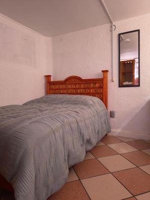 Une chambre avec une tête de lit en bois, un lit recouvert d’une couverture tricotée grise, un sol carrelé et un miroir fixé au mur.