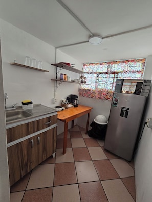 A compact kitchen with a sink, wooden cabinets, a countertop, a table, a refrigerator, and a window with patterned curtains. - Rincon Hortensia apartments and loft - Apt de 1 habitacion 2 (Oaxaca de Juárez)