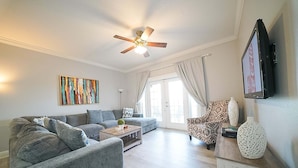 A living room with a sofa, a coffee table, a patterned armchair, a ceiling fan, and a television mounted on the wall. - Sunset Greens Retreat at Reunion-7603 (Kissimmee)